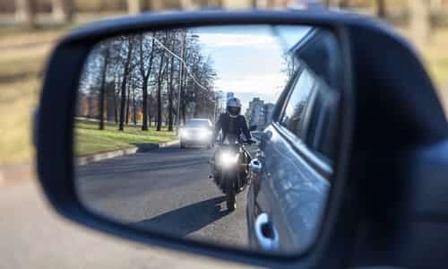 A motorcycle fast approaching a car to pass them as seen from the side mirror.